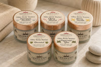 Five Queen City Skincare Silky Body Butter jars in assorted scents arranged on a marble bathroom counter with towels, greenery, and spa ston