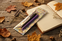 Two pens, black and purple, in a clear pouch resting on an open journal, with autumn leaves and acorns on a wood table.
