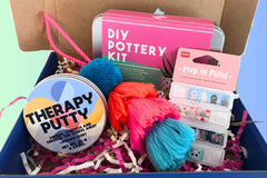A picture of an example box featuring a bright coloured Pompom tassel, DIY pottery kit, therapy putty, a colouring book, page markers & info