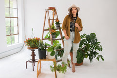 A model in a grunge outfit with heels on and a hat standing in a white room with lots of plants surrounding her. 