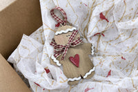 The layered wood gingerbread man ornament up close: white rickrack icing, a red gingham bow and a red heart, in its kraft mailer