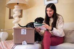 A woman smiles as she reads the booklet from her Discovery Kit. Her dog is with her on the couch and the box is next to them.