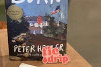 Burn by Peter Heller, a hardcover book, standing upright on a borwn paper bag with a pink and red FicDrip sticker propped in front.