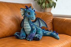 A teal and purple dragon plush rests on a warm leather sofa, showing the whimsical character styles available in our pre-owned plush collect
