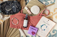 These Palm People box products are displayed on a beach towel: shower steamer, bracelet, small bag, tanner, perfume, and face mask.