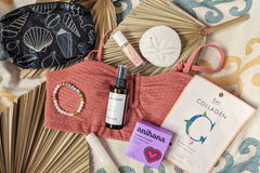 These Palm People box products are displayed on a beach towel: shower steamer, bracelet, small bag, tanner, perfume, and face mask.
