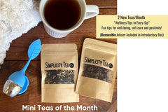 Two kraft paper pouches of Simplicity Tea on a wooden table, next to a cup of tea and a blue leaf-shaped tea infuser. 2 new teas every month