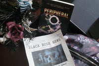 In a black box, a book called Peripheral, the Black Rose newsletter, and a skull and red rose purse. A burgundy rose plant is pictured.