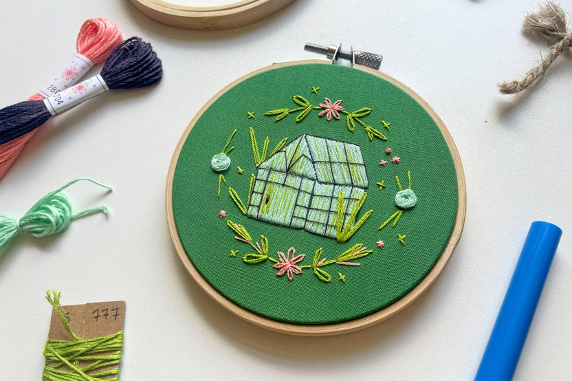 Embroidery project featuring a colorful greenhouse and flowers surrounded by embroidery kit supplies