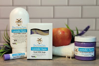 Lavender Apple goat milk soap, lotion, and sugar scrub sits next to a Lavender Vanilla Lip Butter. A real apple and lavender spring are in t