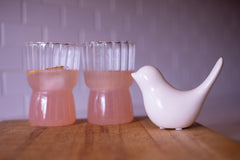 Cute glass mugs with pink lemonade, lemon slices, and a white bird on a wooden cutting board and subway tile background