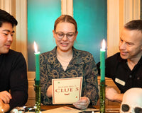 Three people sitting at a table looking at clues of a mystery escape game. With candles and a skeleton next to them.