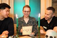 Three people sitting at a table looking at clues of a mystery escape game. With candles and a skeleton next to them.