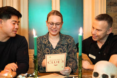 Three people sitting at a table looking at clues of a mystery escape game. With candles and a skeleton next to them.