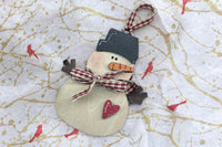 The layered wood snowman ornament up close: a charcoal top hat, carved carrot nose, gingham scarf, twig arms and a red heart