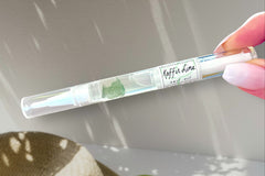 Hand Holding The Kaffir Lime Cuticle Oil Pen by Greenmade goods in Sunlight With a Woven Basket in the Background.