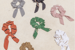 a flat lay of 7 scrunchies of assorted colors laying on the ground, which is tan sandy color, each with a tail