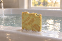 The unwrapped Turmeric & Black Pepper tallow bar, wet with a little lather, resting on a bathtub rim as the bath fills