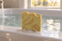 The unwrapped Turmeric & Black Pepper tallow bar, wet with a little lather, resting on a bathtub rim as the bath fills