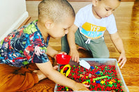 2 small children playing together in a red and green dyed chickpea sensory bin. Children are scooping and sifting through the sensory filler