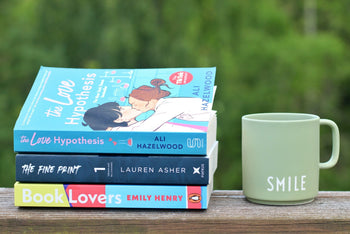 Romance books stacked neatly next to a Smile mug, creating a warm outside, inviting scene for a relaxed reading break.