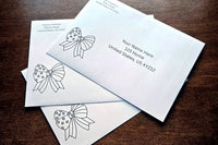 Three white envelopes with printed sample U.S. mailing addresses and black outline patriotic bow designs arranged on a wooden table.