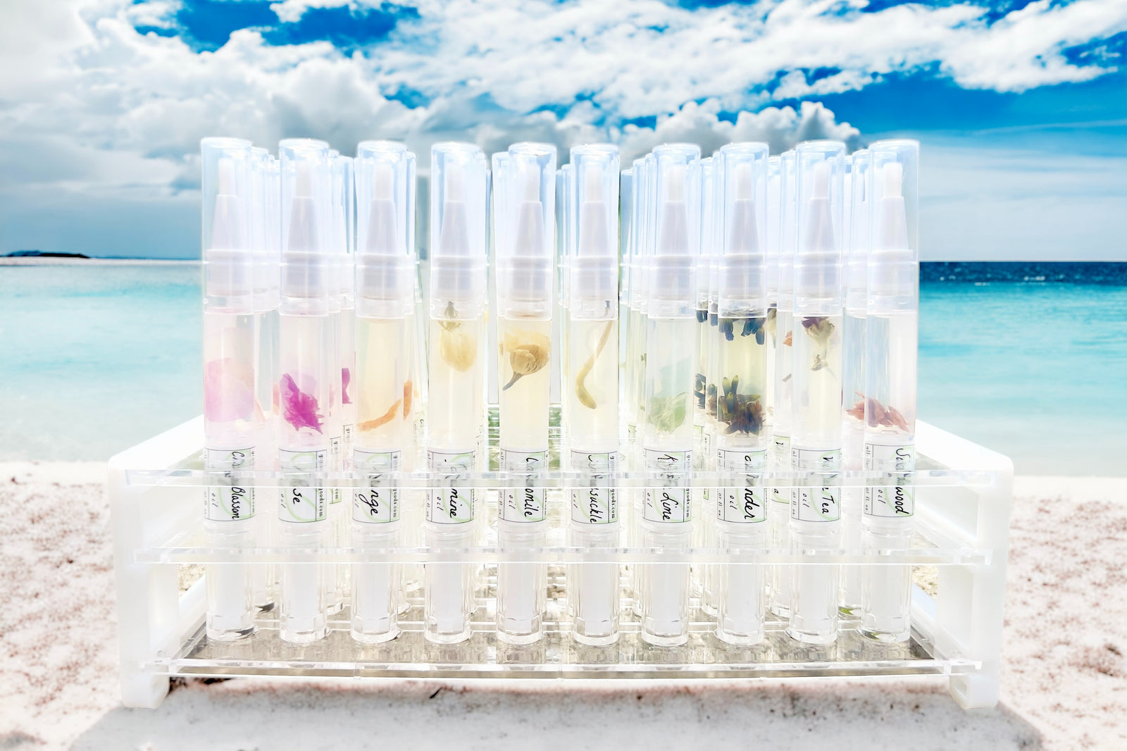 Ten different Greenmade goods cuticle oils in a clear display stand on a sandy white beach with a blue sky and clouds in the background,.