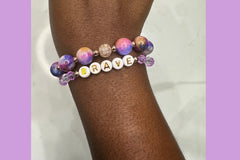 Handmade encouragement bracelet with vibrant purple stone beads, lavender faux crystal accents, and white letter beads spelling “BRAVE” 