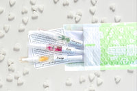 Greenmade goods Lemongrass, Rose, and Orange cuticle oils in a green and white envelope against a white background surrounded by hearts.