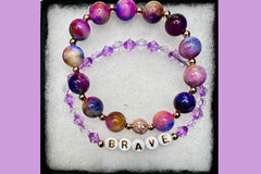 Handmade encouragement bracelet with vibrant purple stone beads, lavender faux crystal accents, and white letter beads spelling “BRAVE” 
