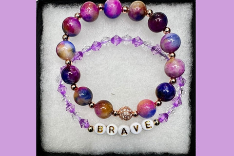 Handmade encouragement bracelet with vibrant purple stone beads, lavender faux crystal accents, and white letter beads spelling “BRAVE” 