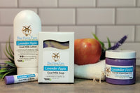 Lavender Apple goat milk lotion, soap, and sugar scrub sits next to a Lavender Vanilla Lip Butter. A real apple and lavender sprig is in the