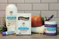 Lavender Apple goat milk lotion, soap, and sugar scrub sits next to a Lavender Vanilla Lip Butter. A real apple and lavender sprig is in the
