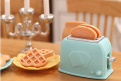 This is a photograph of a tiny blue miniature toaster oven with toast popping out the top.  There are two waffles on a plate.