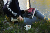 A woman with a sweater pouring liquid to a cup while holding a book called the divergent while sitting next to a lake or river.