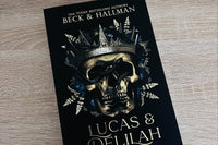 A dark color cover with a golden skull wearing a crown. The title Lucas and Delilah appearing on the bottom with gold lettering. 