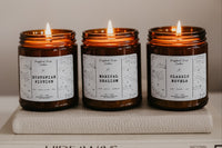 a trio of candles sits atop a stack of books. the candles are lit. The candle names are Dystopian Fiction, Magical Realism, and CLassicNovel