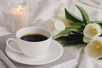 A CUP OF COFFEE SITS ON A SAUCER AND BOTH SIT ATOP A SHORT STACK OF BOOKS. WHITE TULIPS ARE BUNDLED NEARBY. A CANDLE BURNS IN THE BACKGROUND
