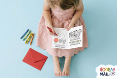 Girl age 2-5 in a light pink dress with her monthly Hooray Mail card open in her lap coloring with a red crayon.