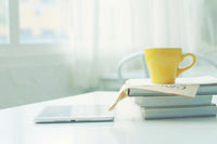 A WHITE TABLE HOLDS A STACK OF WHITE BOOKS. a YELLOW COFFEE CUP SITS STOP THE STACK OF BOOKS. A LAPTOP SITS NEARBY.