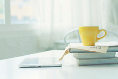 A WHITE TABLE HOLDS A STACK OF WHITE BOOKS. a YELLOW COFFEE CUP SITS STOP THE STACK OF BOOKS. A LAPTOP SITS NEARBY.