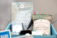 A sample box with Calligraphy book and pens and self care items including 20 minute candles, a crystal fidget, and heated wheat bag