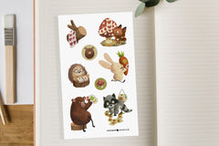 An open notebook with a sticker sheet displayed containing woodland animals, a bear, fox, raccoon etc playing with rolls of stickers.
