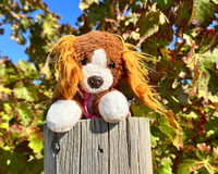 Snorri Stuffed animal, a brown and white dog stuffed animal with a pink bandana on a wooden fence in front of grape leaves. 