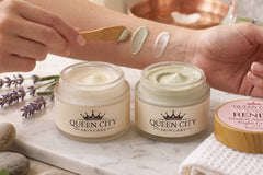 Close-up of cream textures being tested on an arm beside two open Queen City Skincare facial cream jars in a warm spa setting.