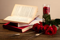 A high quality photo o a oneped book laying on 2 more books with glass with rose in it next to them and also 2 more roses on the table.