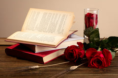 A high quality photo o a oneped book laying on 2 more books with glass with rose in it next to them and also 2 more roses on the table.