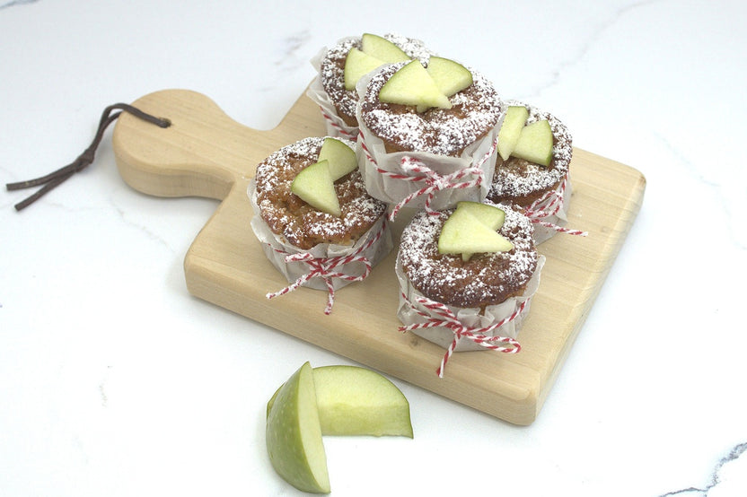 Mini golden apple cakes studded with apple bits, dusted with sugar, alongside muffin pan and baking tools
