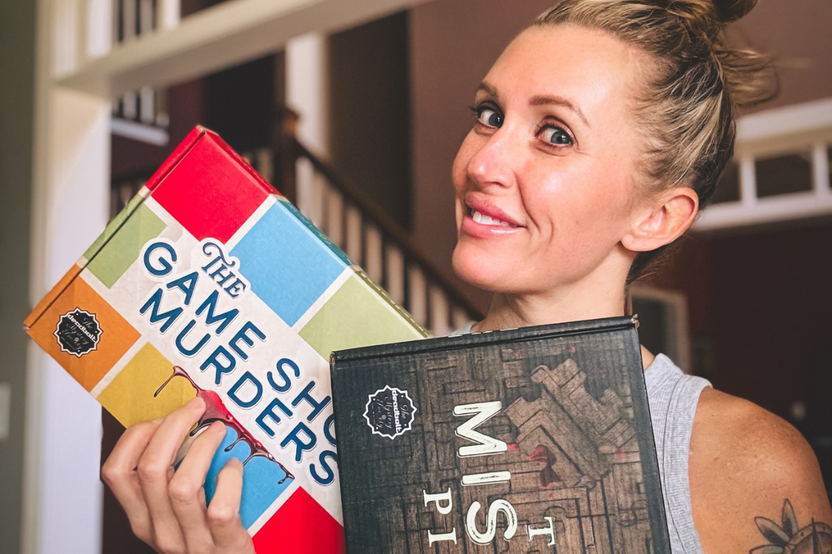A woman holding in her hands two different murder mystery game boxes from The Deadbolt Mystery Society.