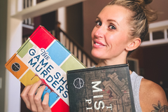 A woman holding in her hands two different murder mystery game boxes from The Deadbolt Mystery Society.